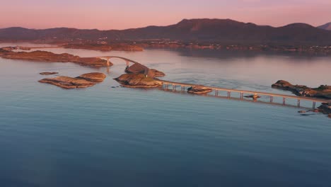 Aerial-shot-of-the-series-of-bridges-of-the-Atlantic-road-stretching-across-islands,-with-a-breathtaking-sunset-and-towering-mountains-as-the-backdrop