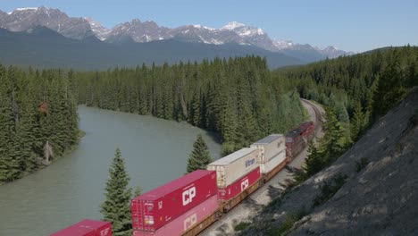 Tren-De-Carga-Viajando-En-Un-Bosque-Con-Montañas-Y-Un-Río-En-El-Parque-Nacional-Banff