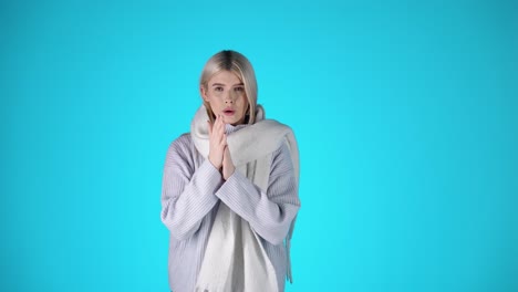 Portrait-Of-Pretty-Blonde-Woman-Feeling-Cold-Shivering,-Warmly-Dressed-Against-Blue-Background,-Studio-Shot