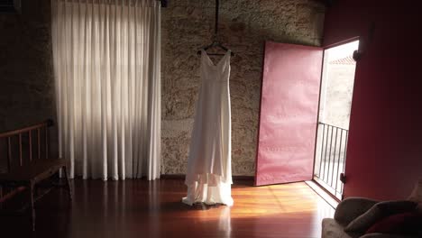 Simple-wedding-dress-hanging-in-a-warmly-lit-room-with-rustic-stone-walls-and-wooden-floors