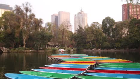 Bunte,-Leere-Ruderboote-Schwimmen-Friedlich-Auf-Einem-See-Vor-Der-Kulisse-Städtischer-Gebäude-Und-üppiger-Vegetation-In-Chengdu,-China.