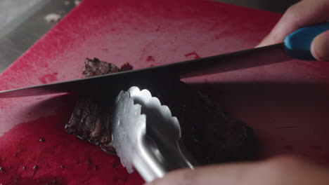 Beautiful-slow-motion,-close-up-shot-of-a-piece-of-cooked-and-flamed-meat-being-cut-into-strips-with-a-large-knife-on-a-red-chopping-board-in-an-industrial-kitchen-in-a-restaurant