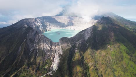 Vista-Aérea-Que-Rodea-El-Borde-De-Un-Volcán-Humeante-Ijen-Con-Un-Lago-Turquesa-Y-Una-Montaña-Nublada-Y-Brumosa-Al-Fondo---Java-Oriental,-Indonesia