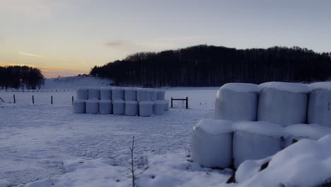 Campo-Agrícola-Cubierto-De-Nieve-Con-Pilas-De-Alimento-Al-Atardecer-Envuelto-En-Nieve