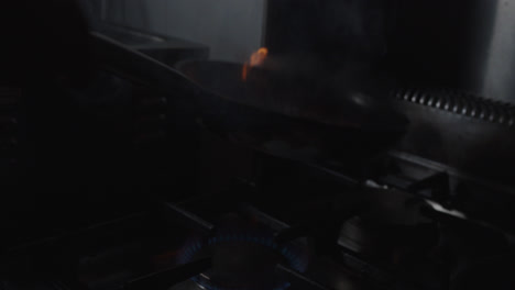 Beautiful-slow-motion-shot-of-a-chef's-culinary-art-while-flaming-a-piece-of-meat-with-a-frying-pan-with-lots-of-fire-in-a-dimly-lit-industrial-kitchen-in-a-restaurant