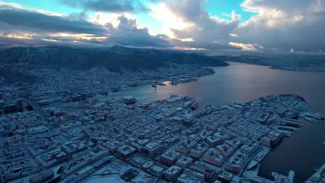 Bergen-establisher-aerial-shot,-Norwegian-town-covered-with-snow-at-sunset,-harbor-fjord