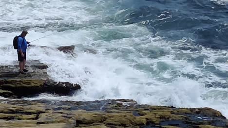 Man-with-hat-and-blue-shirt-standing-on-rocks-very-close-to-the-ocean-in-Two-Lights-State-Park