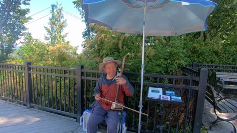 Mann-Mit-Hut-Auf-Dem-Kopf-Sitzt-Auf-Der-Straße-Und-Spielt-Auf-Einem-Musikinstrument