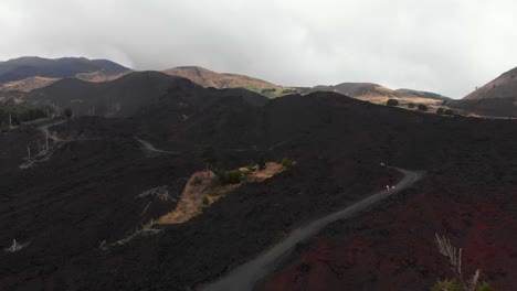 AERIAL-DOLLY-OUT:-Drone-flying-over-a-valley-of-lava-rocks,-in-Mount-Etna,-Sicily,-on-a-foggy-day,-revealing-a-desolate-and-martian-look-panorama-with-the-top-of-the-crater-in-the-distance