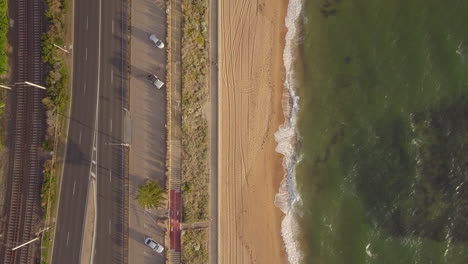 Drone-aerial-top-down-view-of-sea,-beach,-road-and-train-tracks-at-St-Kilda-beach,-cam-moving-right-to-left,-Australia-Melbourne-day-time-sun-setting