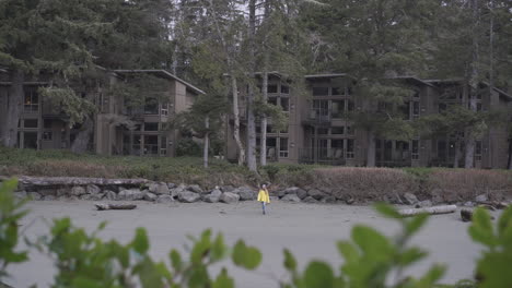 Ein-Mädchen-In-Einem-Gelben-Regenmantel-Spaziert-Am-Strand-Entlang,-Im-Hintergrund-Die-Strandhotels-In-Tofino,-British-Columbia,-Kanada