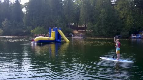 Mit-Der-Luftkamera-Nach-Vorn-Beobachtet-Ein-Junge-Von-Einem-Schwimmenden-Paddleboard-Auf-Einem-See-Aus,-Wie-Andere-Kinder-Von-Einem-Aufblasbaren-„Blob“-Spielzeug-Springen-Und-Ins-Wasser-Fallen