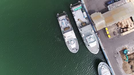 Fishing-Boats-Moored-at-Cargo-Dock-Building,-Green-Sea,-Crates,-ABOVE