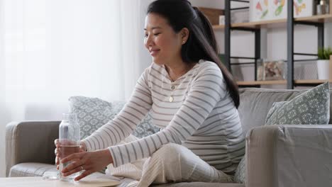 Pregnant-Woman-with-Water-in-Glass-Bottle-at-Home.pregnancy,-rest,-people-and-expectation-concept--happy-smiling-pregnant-asian-woman-sitting-on-sofa-at-home-and-drinking-water-from-reusable-glass-bottle