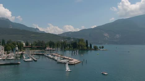 Beautiful-drone-shot-of-the-marina-in-Riva-Del-Garda-on-the-garda-lake-with-a-sailing-boat-in-the-foreground-and-the-italian-alps-in-the-background