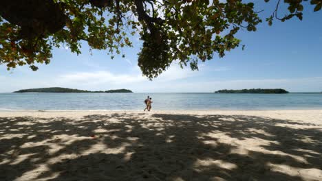 Weitwinkelaufnahme-In-Thailand-Unter-Einem-Baum-Mit-Blick-Auf-Den-Strand-Und-Das-Wasser,-An-Dem-Ein-Paar-Vorbeigeht