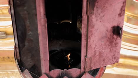 Small-candle-burning-inside-old-rusty-candle-holder-in-front-of-gold-plated-chedi-at-a-Buddhist-temple-in-Chiang-Mai,-Thailand