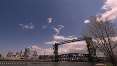 Cleveland,-Ohio,-Time-lapse-Del-Río-Cuyahoga-Con-Un-Puente-De-Tren-En-Primer-Plano-Mientras-Las-Nubes-Pasan-Por-Whiskey-Island-En-Wendy-Park-En-La-Costa-Del-Lago-Erie