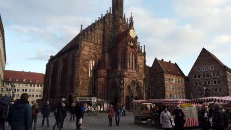 Die-Historische-Frauenkirche-In-Nürnberg-Leuchtet-Im-Abendlicht-Golden-orange,-Während-Unten-Ein-Bauernmarkt-Mit-Vielen-Menschen-Und-Touristen-Stattfindet