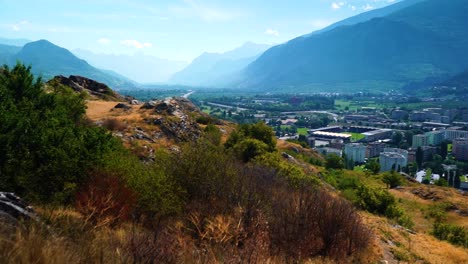 Reveal-shot-of-beautiful-mountains-and-Swiss-valley-city-in-summer-with-plants-and-rocks-foreground