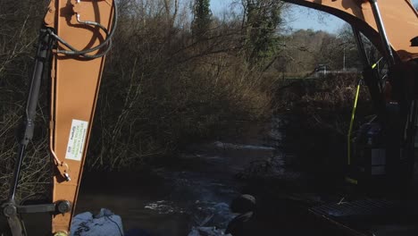 Aerial-flying-backwards-through-the-arm-of-an-excavator-over-a-stream-in-a-wooded-area-in-Autumn