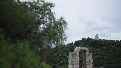 Vista-Lenta-Desde-Detrás-De-Un-árbol-Con-Piñas-Para-Ver-La-Cima-De-La-Colina-Filopappou-En-Un-Día-Nublado-Y-Melancólico-Con-Ruinas-De-Piedra-Que-Emergen-De-Los-árboles.