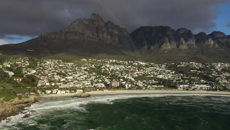 Toma-Aérea-Con-Arcoíris-De-La-Playa-De-Camps-Bay-En-Ciudad-Del-Cabo-Con-La-Montaña-De-La-Mesa-De-Fondo-Al-Atardecer