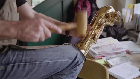 Skilled-Instrument-maker-repairing-dents-in-golden-saxophone-bell-and-bow-with-hammer-on-his-knee