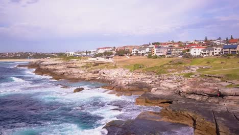 Aerial-shot-of-drone-flying-over-a-rock-pool-near-the-beach-with-ocean-waves-crashing-on-the-rocks-in-the-countryside-beach-front-houses