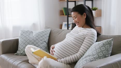 Mujer-Embarazada-Feliz-Leyendo-Un-Libro-En-Casa.-Concepto-De-Embarazo,-Personas-Y-Ocio.-Mujer-Asiática-Embarazada-Feliz-Y-Sonriente-Sentada-En-El-Sofá-Y-Leyendo-Un-Libro-En-Casa