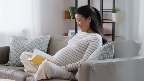 Mujer-Embarazada-Feliz-Leyendo-Un-Libro-En-Casa.-Concepto-De-Embarazo,-Personas-Y-Ocio.-Mujer-Asiática-Embarazada-Feliz-Y-Sonriente-Sentada-En-El-Sofá-Y-Leyendo-Un-Libro-En-Casa