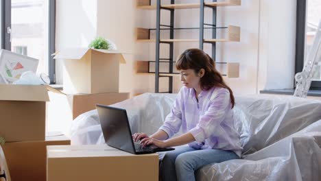 Mujer-Con-Computadora-Portátil-Mudándose-A-Una-Nueva-Casa.-Mudanza,-Personas-Y-Concepto-De-Bienes-Raíces.-Mujer-Asiática-Feliz-Y-Sonriente-Con-Computadora-Portátil-Y-Cajas-En-Una-Nueva-Casa