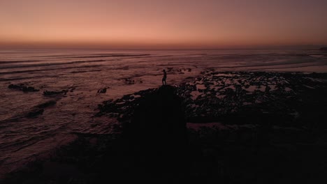 A-aerial-view-of-a-beautiful-sunset-over-a-beach-in-Costa-Rica,-with-waves-and-somebody-on-top-of-a-huge-and-unique-rock