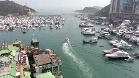 Small-Yacht-leaving-Hong-Kong-marina-and-Typhoon-shelter-at-Po-Chong-Wan-bay-with-skyscrapers-and-hundreds-of-small-boats-on-a-clear-Summer-day,-Aerial-view