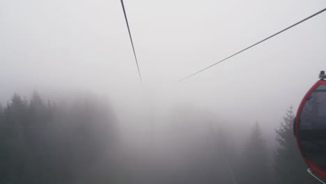 Red-cable-car-moving-away,-very-foggy-view-above-the-trees-in-Auffach,-Austria