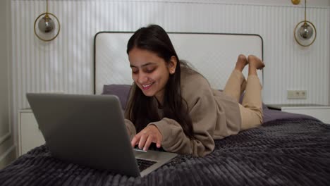 Ein-Glückliches-Brünettes-Mädchen-In-Einem-Braunen-Pullover-Liegt-Auf-Dem-Bett-Und-Tippt-Auf-Einem-Grauen-Laptop,-Während-Sie-In-Einem-Modernen-Apartment-Im-Schlafzimmer-Arbeitet-Und-Zeit-Online-Verbringt