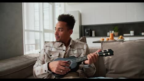 A-calm-and-happy-man-with-Black-skin-color-a-young-brunette-with-stubble-plays-a-small-musical-stringed-instrument-the-ukulele-while-sitting-on-a-modern-sofa-in-a-cream-plaid-shirt-in-a-modern-apartment