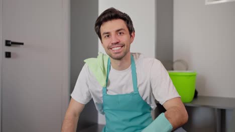 Porträt-Eines-Selbstbewussten,-Glücklichen-Brünetten-Reinigungsmannes-In-Blauer-Schürze-Und-Weißem-T-Shirt,-Der-Jetzt-In-Einem-Modernen-Apartment-Für-Eine-Reinigungsfirma-Auf-Abruf-Arbeitet