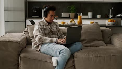 Confident-and-happy-man-with-Black-brunette-skin-color-in-black-modern-headphones-plays-video-games-on-his-old-laptop-while-sitting-on-a-modern-sofa-in-a-studio-apartment