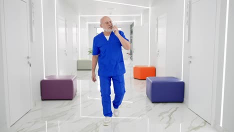 A-confident-head-physician-with-a-gray-beard-a-bald-man-in-a-blue-uniform-quickly-walks-along-the-corridor-and-hurries-while-communicating-on-a-white-phone.-Confident-male-doctor-rushes-to-a-call-along-the-corridor-in-a-modern-clinic