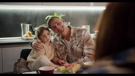 Over-the-shoulder-portrait-of-a-happy-middle-aged-man-with-gray-hair-along-with-his-little-daughter-with-brown-hair-with-a-braided-hairstyle-who-are-sitting-at-the-table-and-chatting-together-with-a-brunette-woman-during-a-family-dinner-in-the-evening