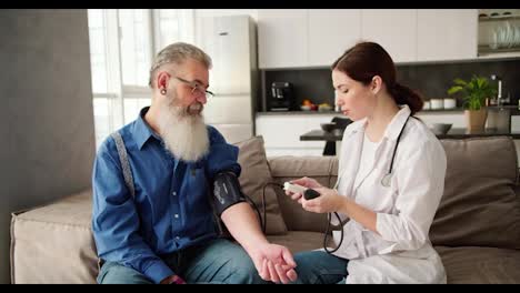 A-brunette-woman-in-a-white-coat-a-doctor-expresses-her-opinion-about-the-pulse-after-examining-an-elderly-man-with-gray-hair-and-a-lush-beard-in-glasses-in-a-blue-shirt-at-home-on-the-sofa-and-they-are-happy