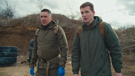 Two-confident-military-men-in-green-uniforms-and-with-backpacks-on-their-backs-walk-along-a-combat-training-ground-with-a-barricade-of-car-tires-and-communicate-in-the-steppe