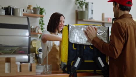 Confident-brunette-girl-puts-an-order-in-a-cardboard-box-for-a-delivery-guy-with-a-big-yellow-bag-in-a-cafe