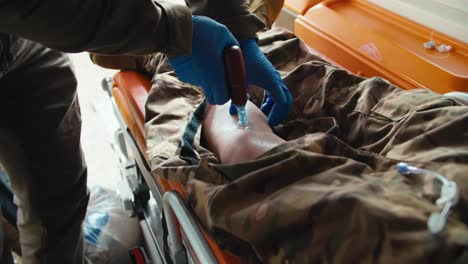 Close-up-of-a-confident-male-army-medic-in-a-dark-green-uniform-and-blue-gloves-inserting-a-needle-into-the-leg-of-an-unconscious-male-soldier-wounded-during-combat-in-the-ambulance-cabin