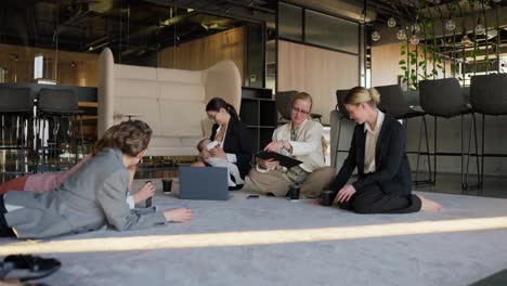 Zoom-in-group-of-confident-businesswomen-in-business-clothes-talking-while-sitting-on-a-gray-sofa-while-a-brunette-businesswoman-girl-breastfeeds-her-small-child