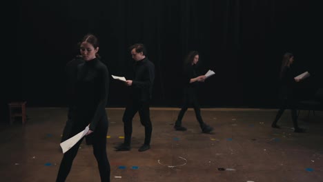 A-group-of-confident-actors-in-black-suits-walk-around-the-stage-with-sheets-of-script-and-text-in-their-hands.-A-brunette-girl-stands-up-straight-and-reads-her-text-before-the-start-of-the-performance-In-the-theatre