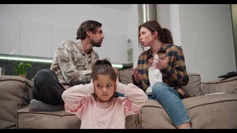 Little-brunette-girl-in-pink-clothes-covers-her-ears-and-is-sad-while-her-parents-are-quarreling-while-sitting-on-the-sofa-in-a-modern-apartment.-A-man-with-stubble-in-a-beige-T-shirt-is-quarreling-with-his-brunette-wife-in-a-plaid-shirt-while-his-little-daughter-is-sitting-on-the-floor-and-covering