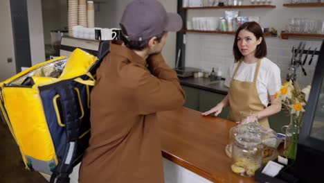 Ein-Mann-In-Einem-Braunen-Pullover,-Der-Essen-Ausliefert,-Kommt-Ins-Café-Und-Spricht-Mit-Dem-Barista,-Während-Er-Die-Bestellung-Bestätigt