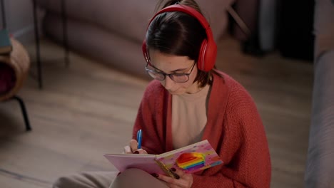 A-confident-brunette-girl-in-red-wireless-headphones-glasses-and-a-red-sweater-sits-on-the-floor-in-a-modern-apartment-and-makes-notes-in-her-notebook